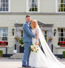 A couple, the bride in a white gown and veil holding a bouquet, and the groom in a dark suit, posing outdoors next to a grand building.