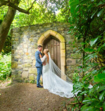 A groom and bride smile while standing outside, surrounded by greenery and trees. The bride holds a bouquet of white and pink flowers.