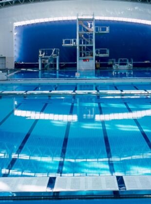 A modern aquatic center with a spacious indoor swimming pool, large windows allowing natural light, and a sleek design featuring wooden ceiling panels and white tiled floors.