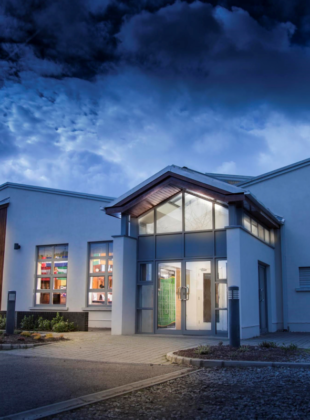 Image depicting a modern clubhouse building with large windows, set against a clear sky. The structure is surrounded by well-maintained greenery, depicting a serene and inviting atmosphere.