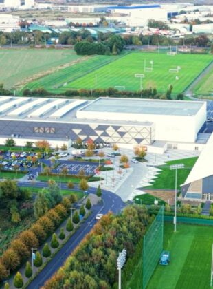 Aerial view of a modern sports campus featuring a swimming pool, tennis courts, sports fields, and surrounding green spaces, with a large building at the center.